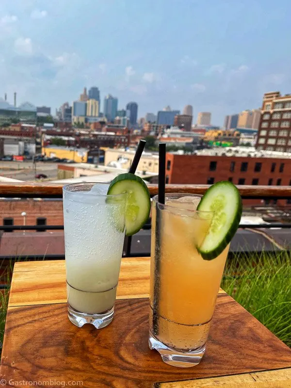 Orange cocktail and opaque cocktail with cucumbers and straws overlook downtown Kansas City