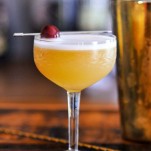 Peach Bourbon Sour, yellow cocktail with white foam in cocktail coupe, cherry on pick. Gold Barware behind