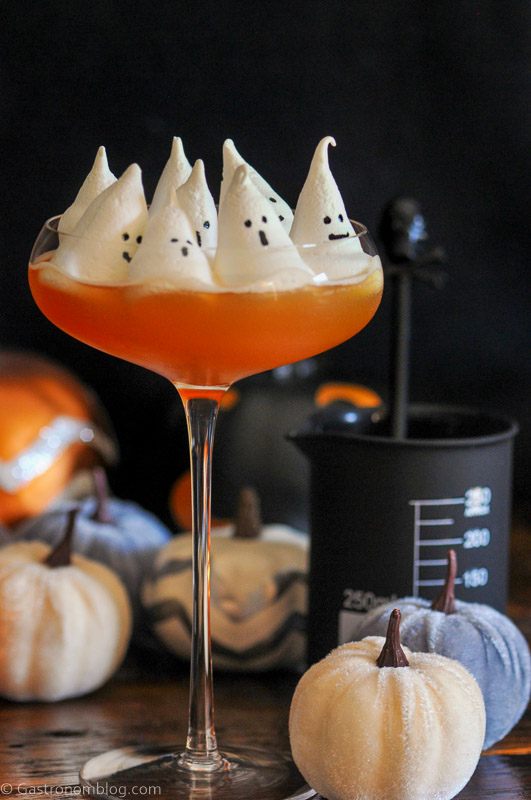 Halloween cocktail recipe, orange in coupe with white ghost meringues on top, pumpkins behind