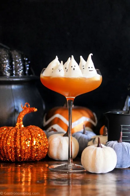 Halloween cocktail recipe, orange in coupe with white ghost meringues on top, pumpkins behind