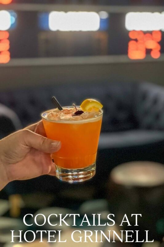 orange cocktail held by hand in front of blue couch and scoreboard