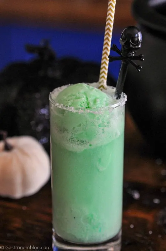 Green Halloween Punch in tall glass with straw and skull stirrer, black cauldron and pumpkins behind