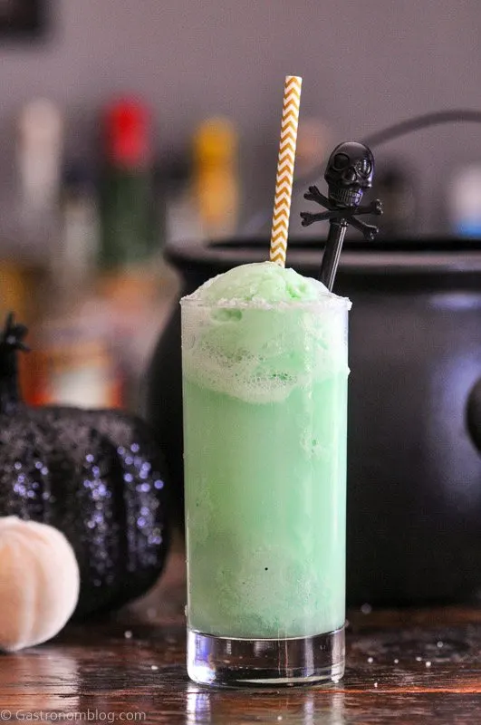Green Halloween Punch in tall glass with straw and skull stirrer, black cauldron and pumpkins behind