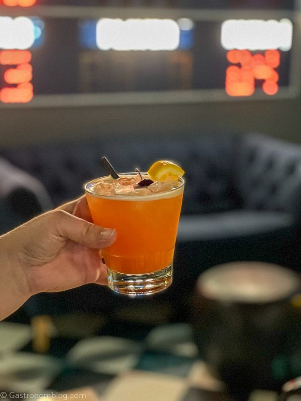 Hand holding orange cocktail in front of scoreboard and blue couch