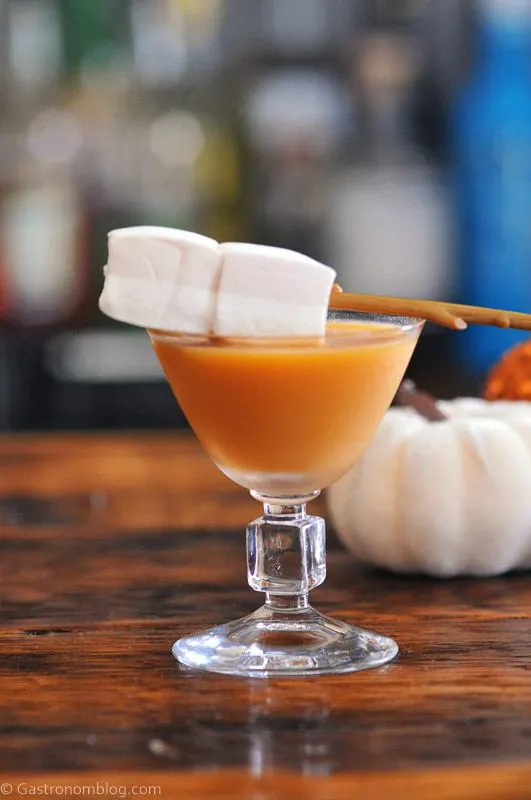 Orange cocktail in glass, marshmallows on stick as garnish. Pumpkins behind
