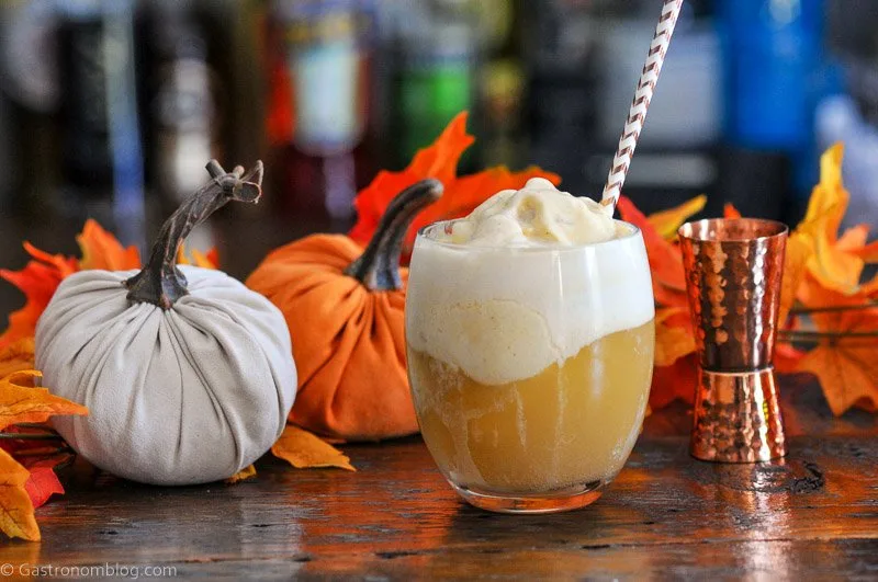 Orange Pumpkin Ice Cream Float in glass, topped with white foam, striped straw, pumpkins and brass jigger