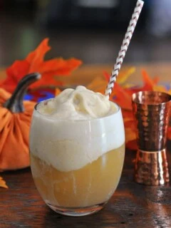 Orange Pumpkin Ice Cream Float in glass, topped with white foam, striped straw, pumpkins and brass jigger