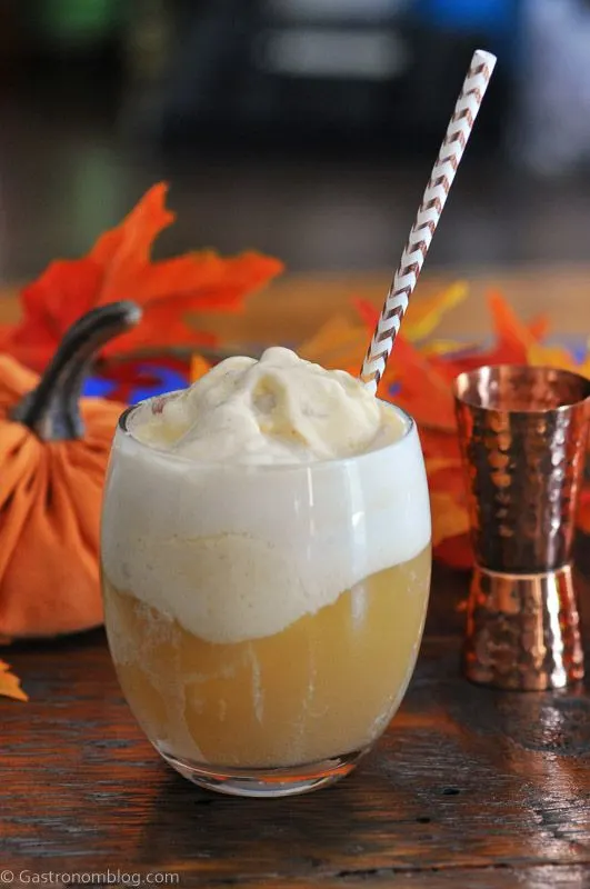 Pumpkin Ice Cream Float in glass, topped with white foam, striped straw, pumpkins and brass jigger