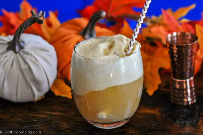 Pumpkin Ice Cream Float in glass, topped with white foam, striped straw, pumpkins and brass jigger