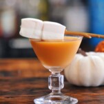 Orange cocktail in glass, marshmallows on stick as garnish. Pumpkins behind