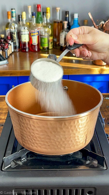 sugar being sprinkled from a measuring cup into copper saucepan