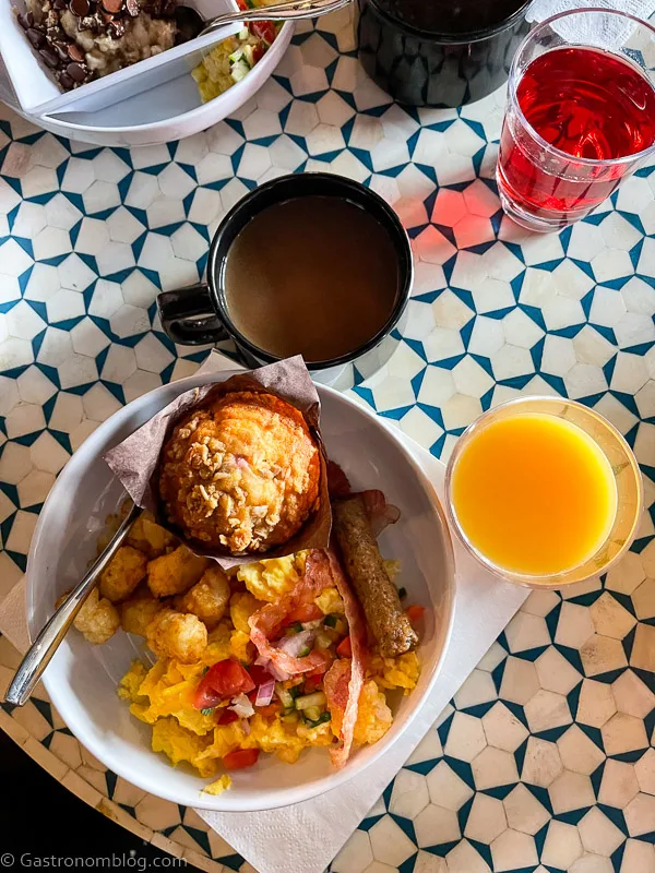 top shot of breadfast - eggs, muffin and potatoes in white bowl with orange juice and coffee on blue and white table