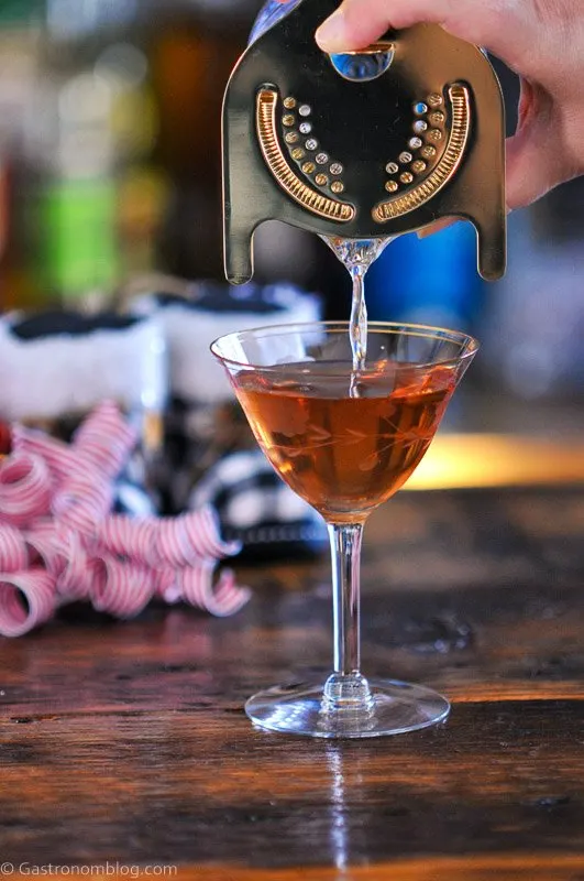 Brown cocktail being poured from mixing glass into coupe