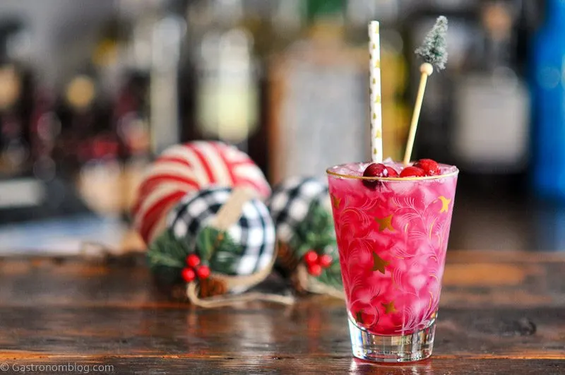 Pink holiday cocktail recipe in gold glass, cranberries and straw, pine tree swizzle stick and Christmas stuff behind