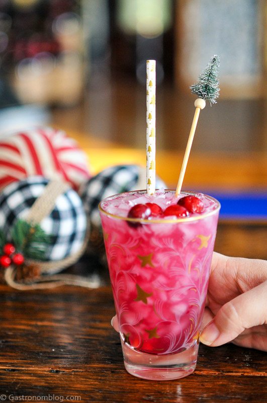 Pink holiday cocktail recipe in gold glass, cranberries and straw, pine tree swizzle stick and Christmas stuff behind
