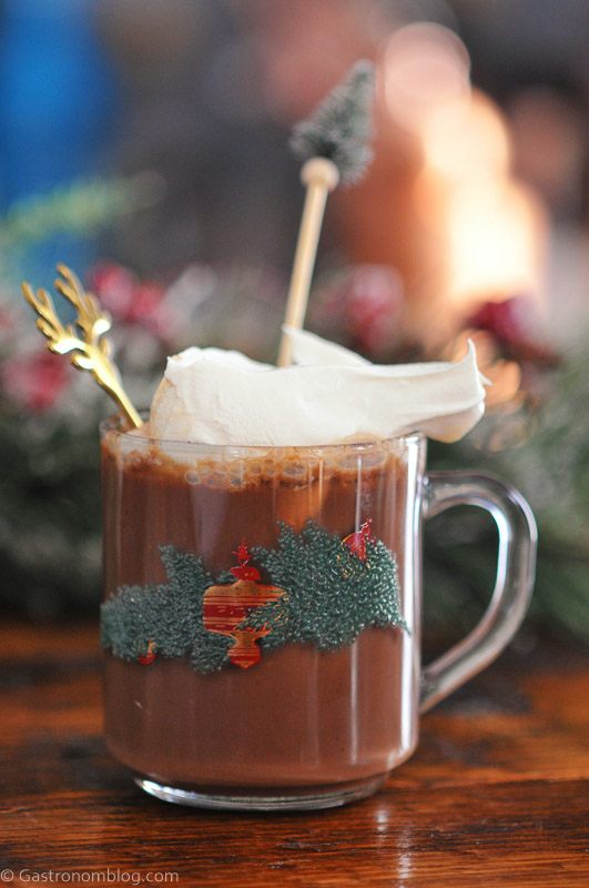 Spiked Hot Chocolate in a glass mug with holly, whipped cream on top, pine stir stick and gold deer spoon