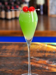 Green cocktail in tall glass, this Grinch cocktail has 2 cherries on a pick as a garnish