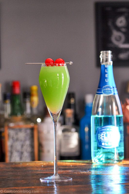 Green cocktail in tall glass, this Grinch cocktail has 2 cherries on a pick as a garnish