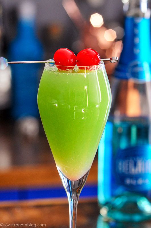 Green cocktail in tall glass, this Grinch cocktail has 2 cherries on a pick as a garnish