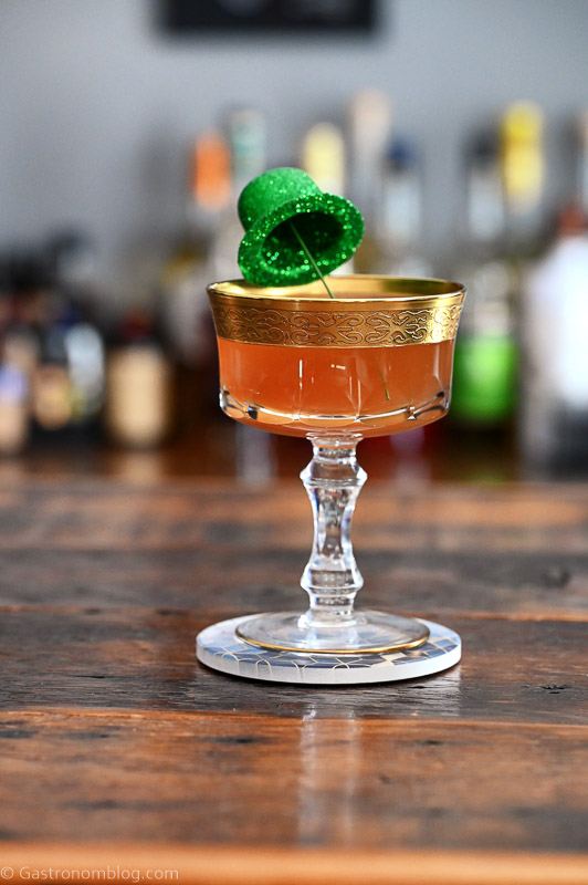 Tan irish Sour cocktail in gold rimmed coupe, green glitter derby hat garnish