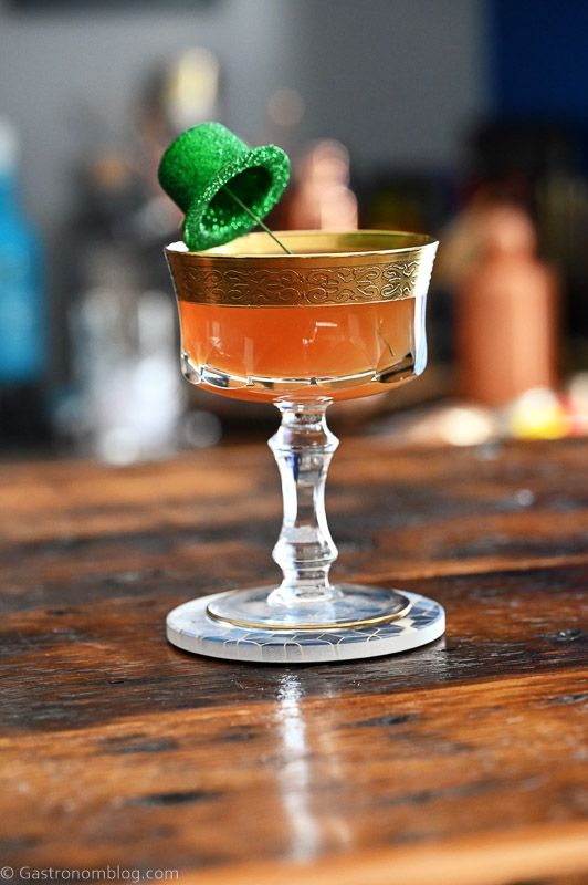Tan irish Sour cocktail in gold rimmed coupe, green glitter derby hat garnish