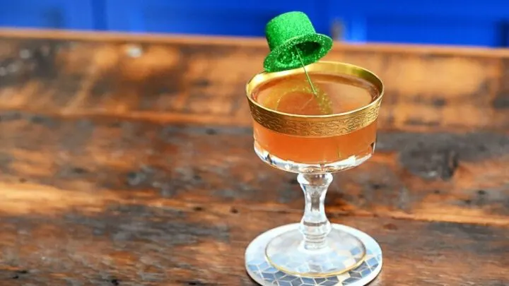 Tan irish Sour cocktail in gold rimmed coupe, green glitter derby hat garnish