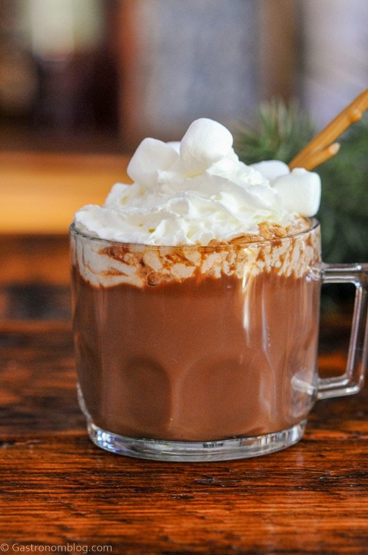 Brown hot chocolate in clear mug, topped with whipped cream and marshmallows. Pine in background.