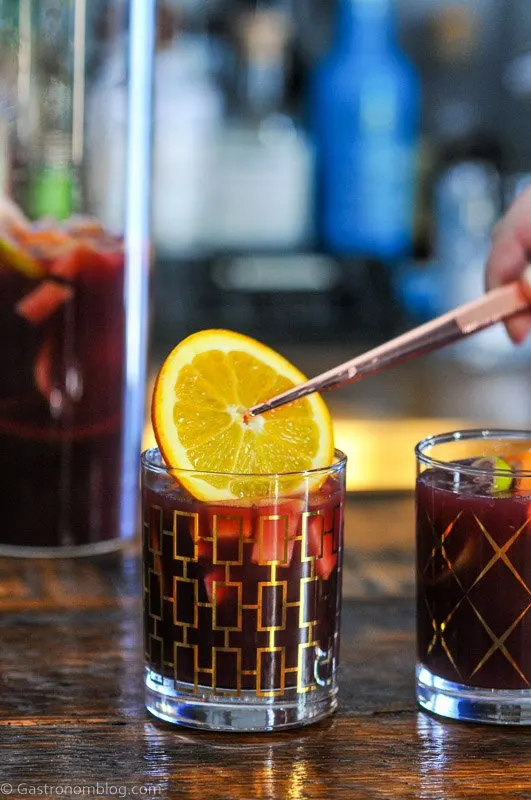 Tongs placing an orange into a gold glass with purple non alcoholic sangria