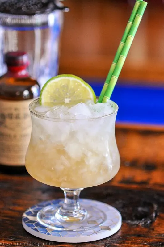 Gold liquid in pebble ice filled glass, bamboo straws, bottle and shaker behind for Winter Mai Tai Cocktail