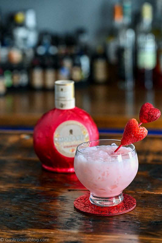 Pink cocktail with strawberry on heart pick, this Strawberry Vodka Cocktail has Strawberry Liqueur bottle behind