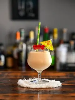 white Bailey's Colada cocktail in glass on white coaster with yellow umbrella, cherry and bamboo straw