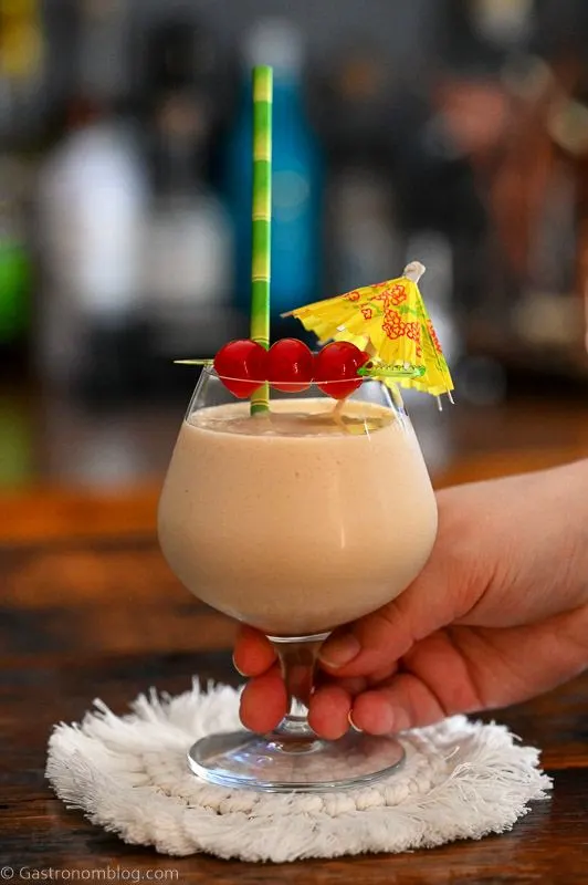 white Bailey's Colada cocktail in glass on white coaster with yellow umbrella, cherry and bamboo straw