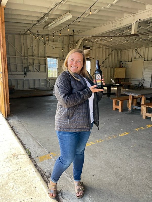 Woman in gray jacket and jeans holding beer bottle at Humboldt Bay Social Club Hangar