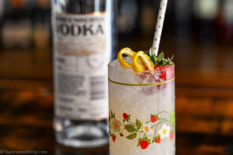Vodka Spritzer cocktail in strawberry print glass with strawberry, lemon peel and straw, vodka bottle behind
