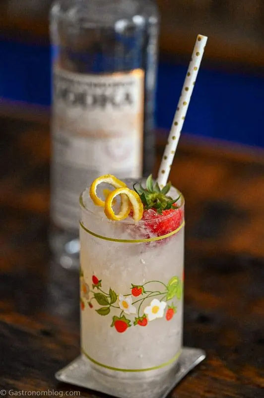 Vodka Spritzer cocktail in strawberry print glass with strawberry, lemon peel and straw, vodka bottle behind