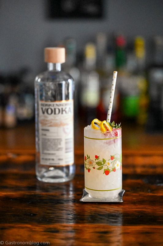 Vodka Spritzer cocktail in strawberry print glass with strawberry, lemon peel and straw, vodka bottle behind