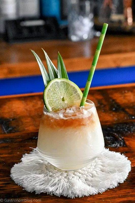 Mai Tai cocktail with dark rum layer on top with lime and pineapple fronds. On white coaster.