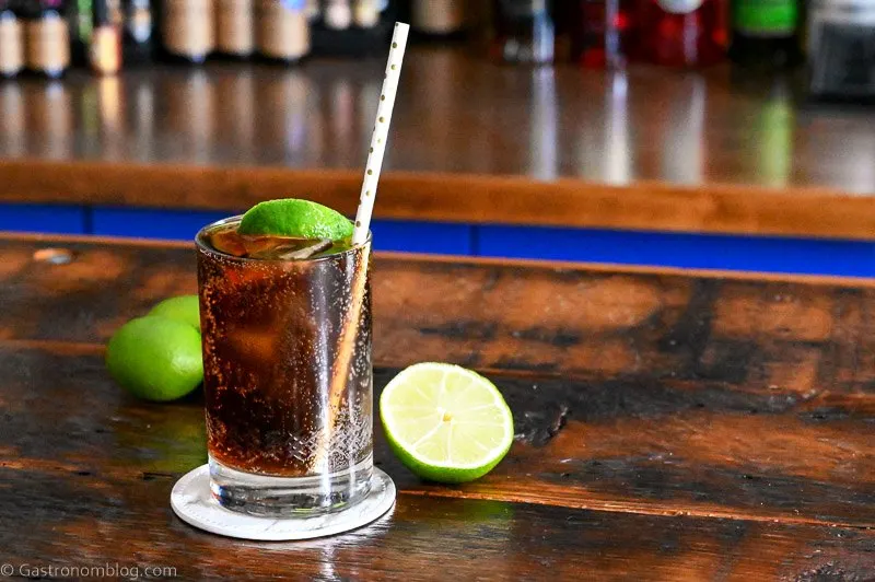 Brown cocktail with ice in a cocktail glass with a straw and lime wedge. Limes behind on wood table.