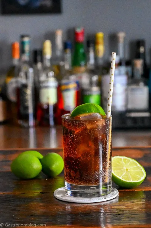 Brown cocktail with ice in a cocktail glass with a straw and lime wedge. Limes behind on wood table.