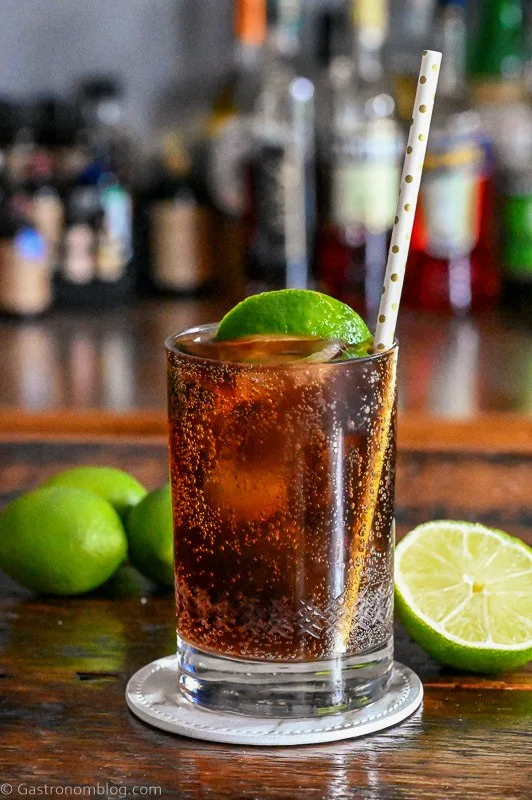 Brown cocktail with ice in a cocktail glass with a straw and lime wedge. Limes behind on wood table.