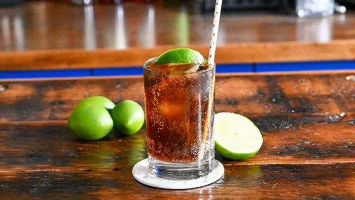 Brown cocktail with ice in a cocktail glass with a straw and lime wedge. Limes behind on wood table.