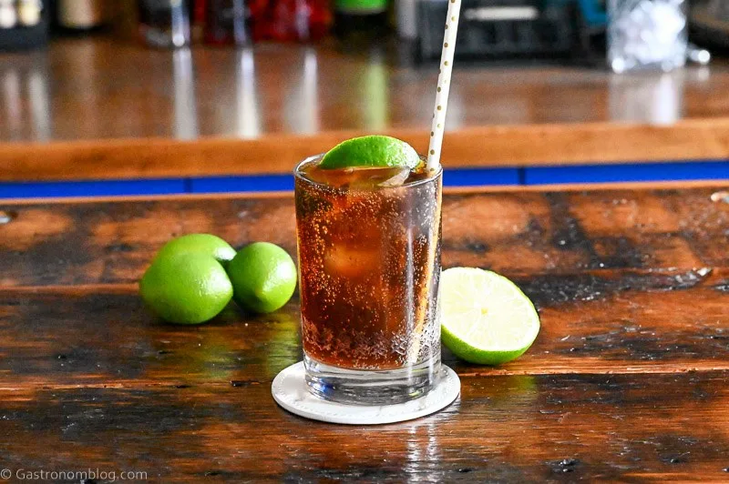 Brown cocktail with ice in a cocktail glass with a straw and lime wedge. Limes behind on wood table.