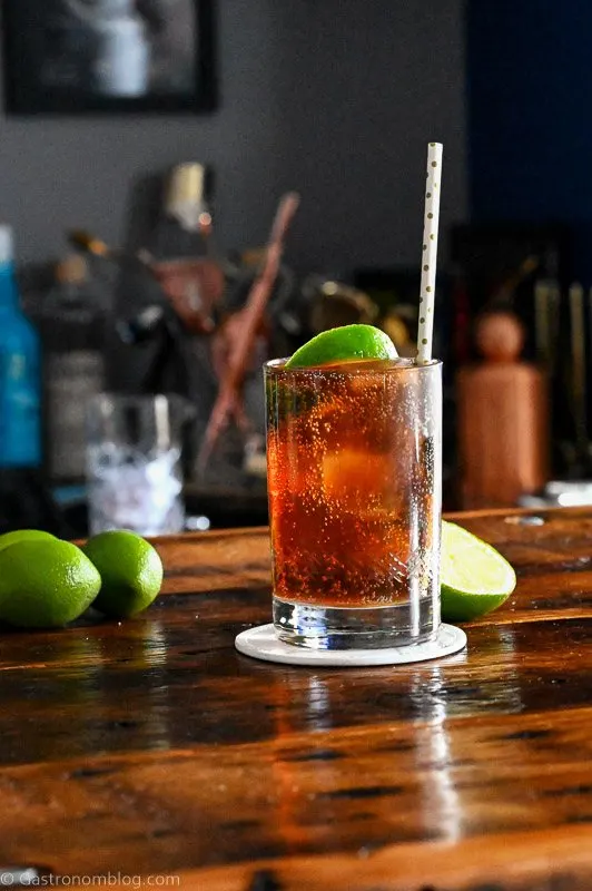 Brown cocktail with ice in a cocktail glass with a straw and lime wedge. Limes behind on wood table.