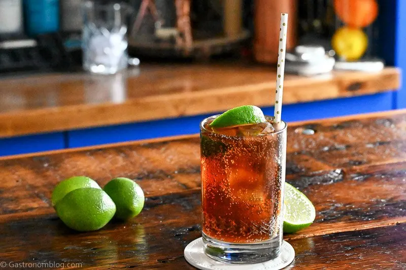 Brown cocktail with ice in a cocktail glass with a straw and lime wedge. Limes behind on wood table.