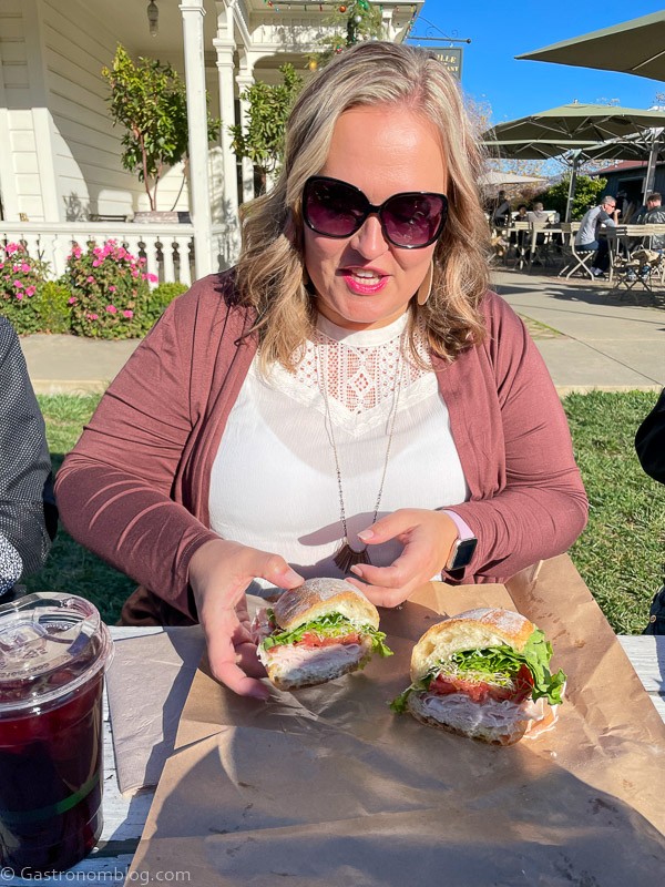 Woman in white shirt and pink cardigan, holding a sandwich outside at Oakville Grocery Napa Valley