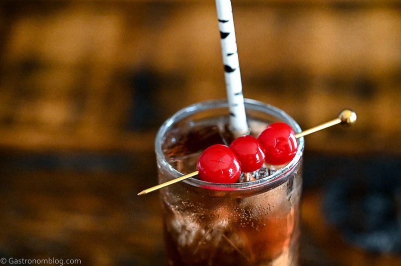 Dirty Shirley Recipe cocktail, a brown cocktail in a tall glass with cherries on a cocktail pick and straw