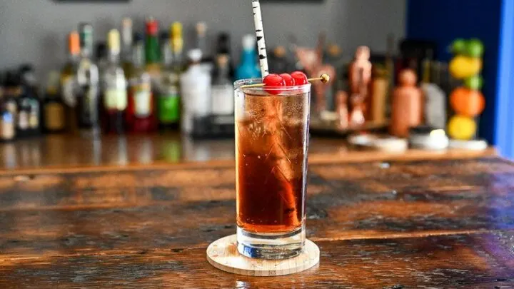 Dirty Shirley Recipe cocktail, a brown cocktail in a tall glass with cherries on a cocktail pick and straw