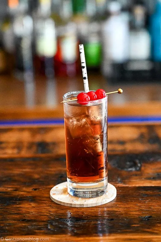 Dirty Shirley Recipe cocktail, a brown cocktail in a tall glass with cherries on a cocktail pick and straw