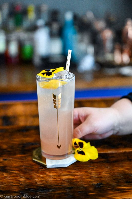 Rhubarb Collins, pink cocktail in tall glass with gold arrow, straw and edible yellow flower, hand holding glass