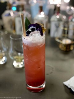 Red cocktail in tall glass with white foam and straw, purple flower garnish on gray bartop
